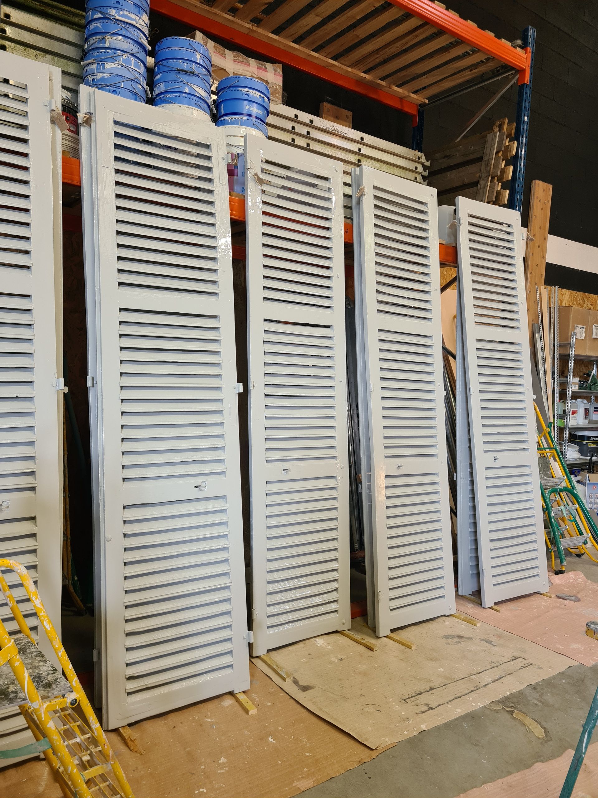 Cinq volets persiennes gris appuyés contre un mur d'un entrepôt ; des seaux bleus sur une étagère en arrière-plan.