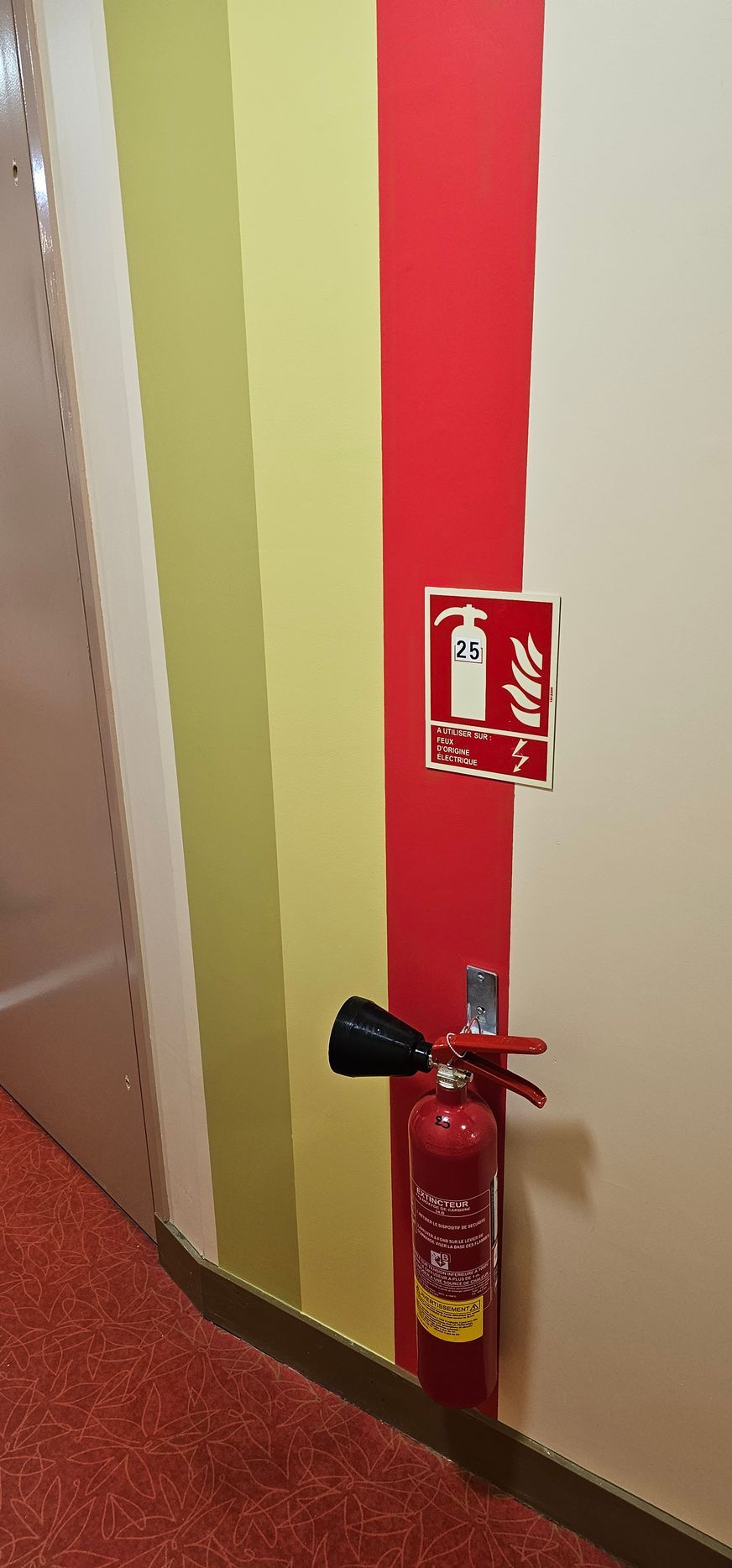 Un extincteur rouge fixé au mur à rayures rouges, jaunes et beiges, avec un panneau au-dessus.