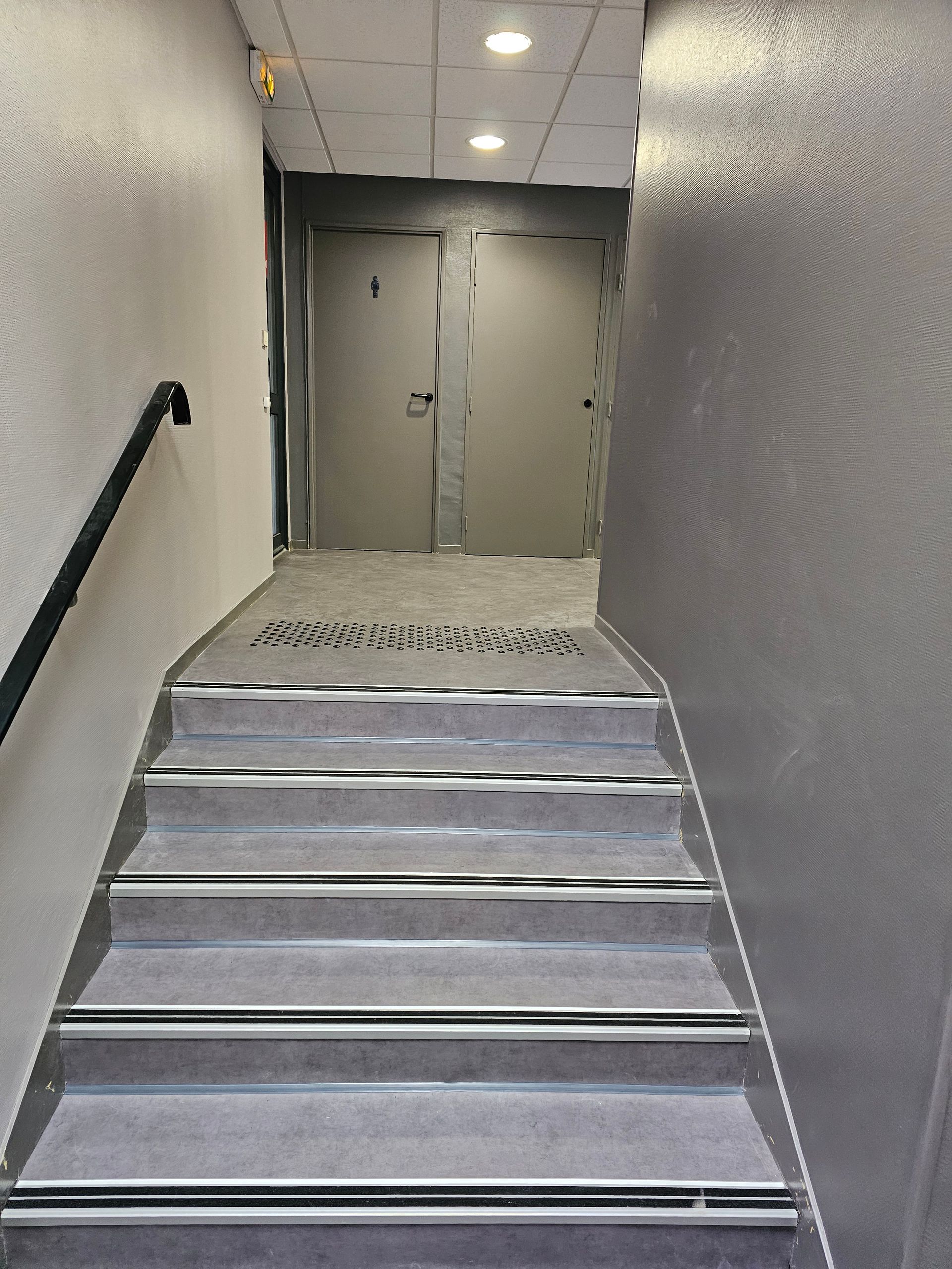 Escalier menant à une porte. Marches grises avec bordures métalliques, murs gris et rampe noire sur la gauche.