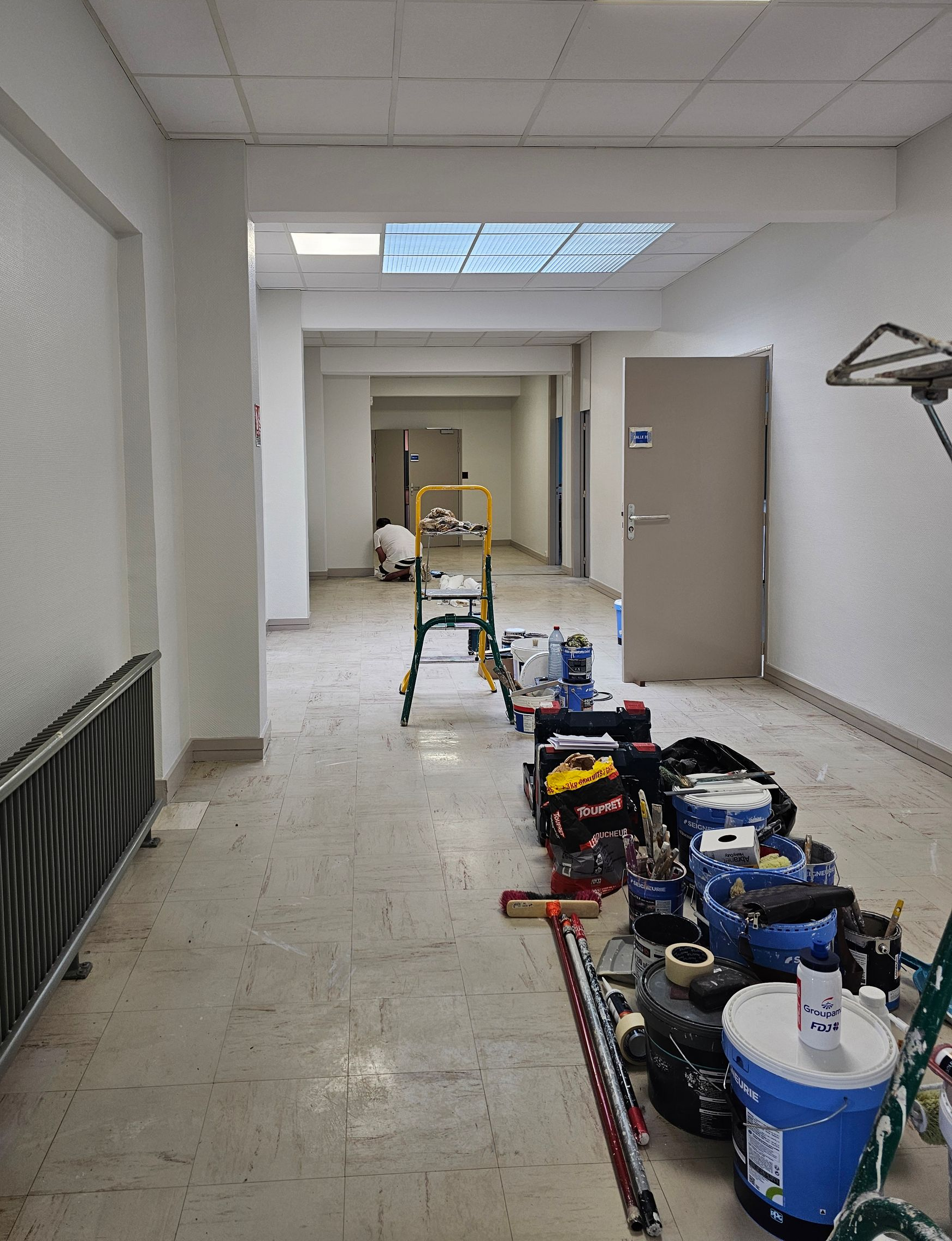 Couloir en rénovation : matériel de peinture, portes et alarme incendie. Murs et plafond blancs.