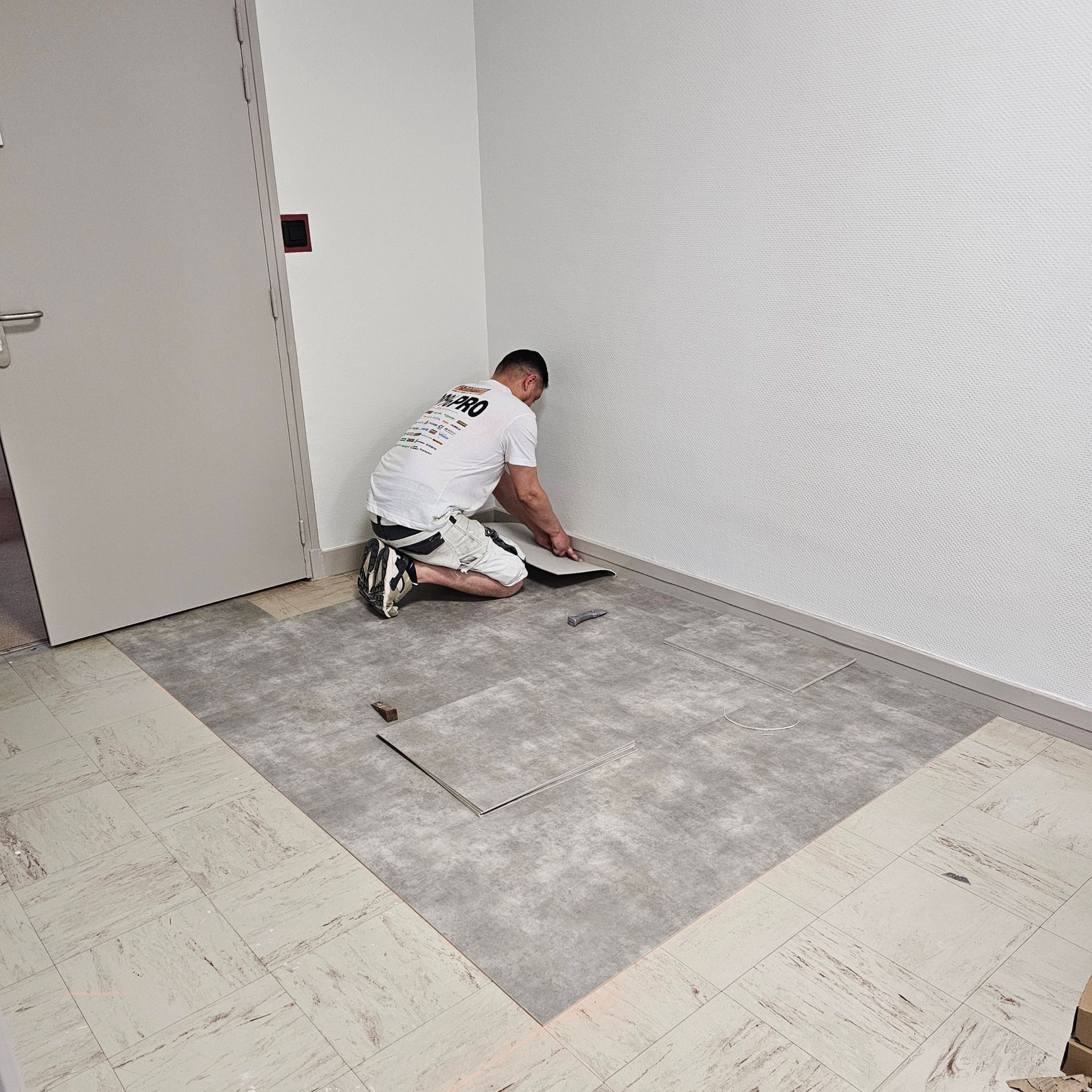 Une personne installe un revêtement de sol gris dans une pièce aux murs et à la porte blancs.