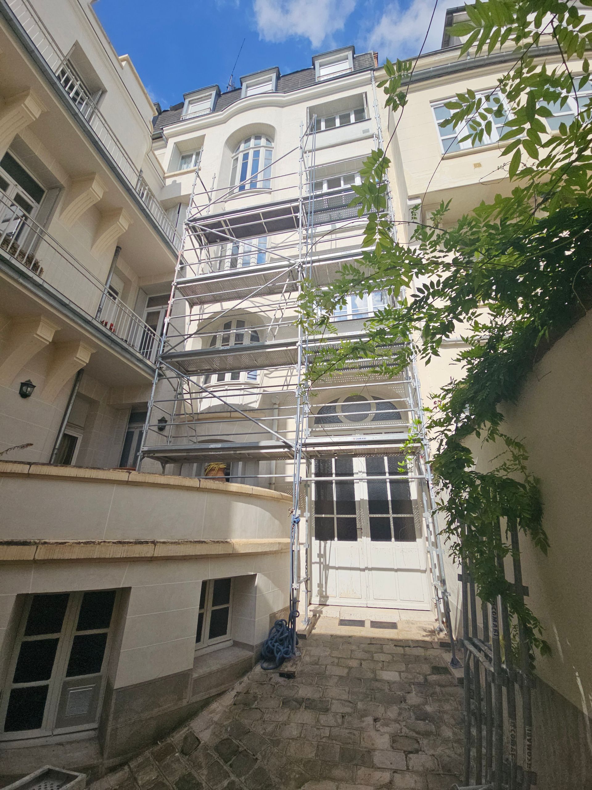Extérieur du bâtiment avec échafaudages pour rénovation. Bâtiments blancs et beiges, porte ouverte au rez-de-chaussée.