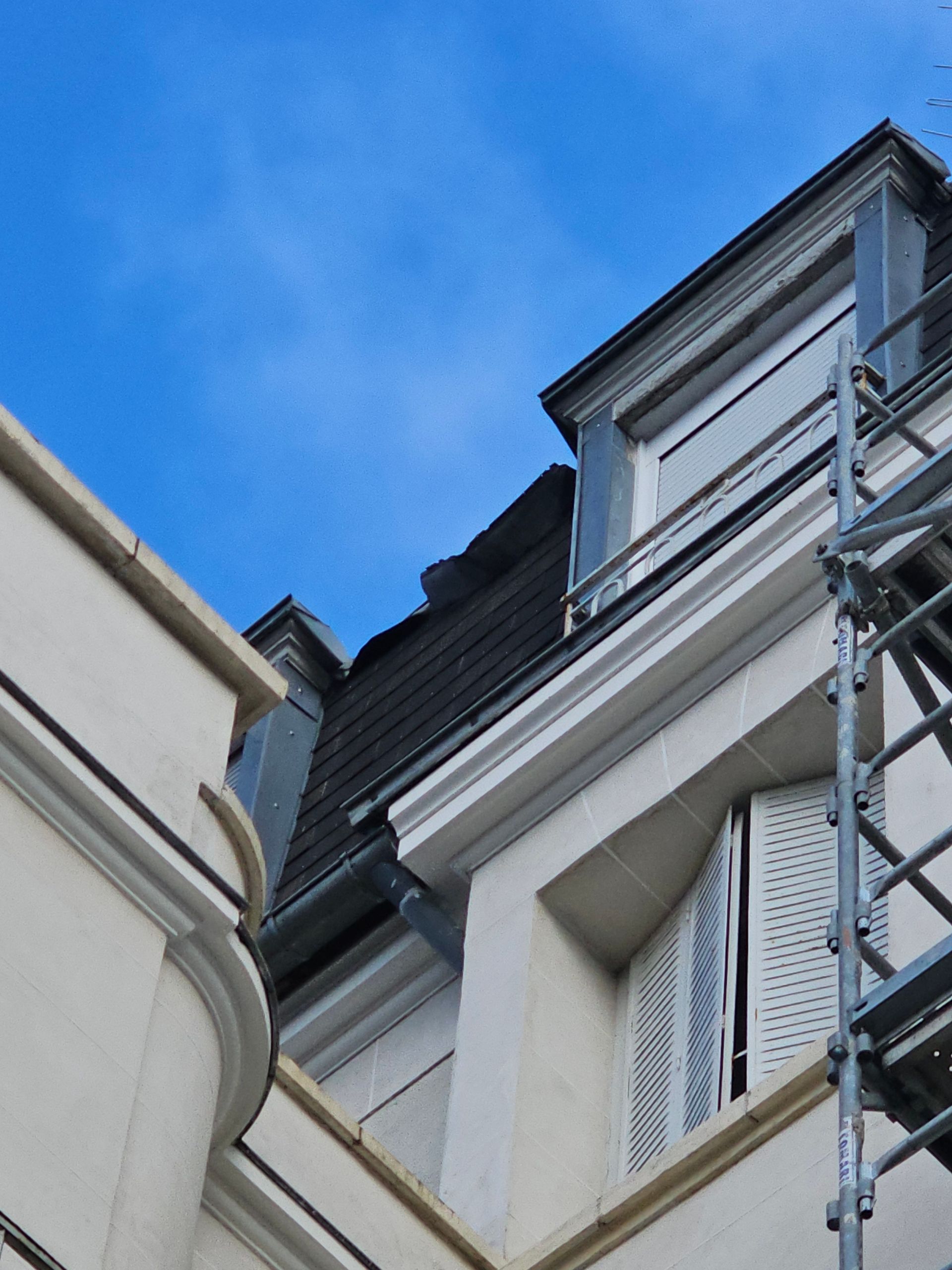 Vue extérieure d'un bâtiment à façade blanche, volets, toit sombre et échafaudages sous un ciel bleu.