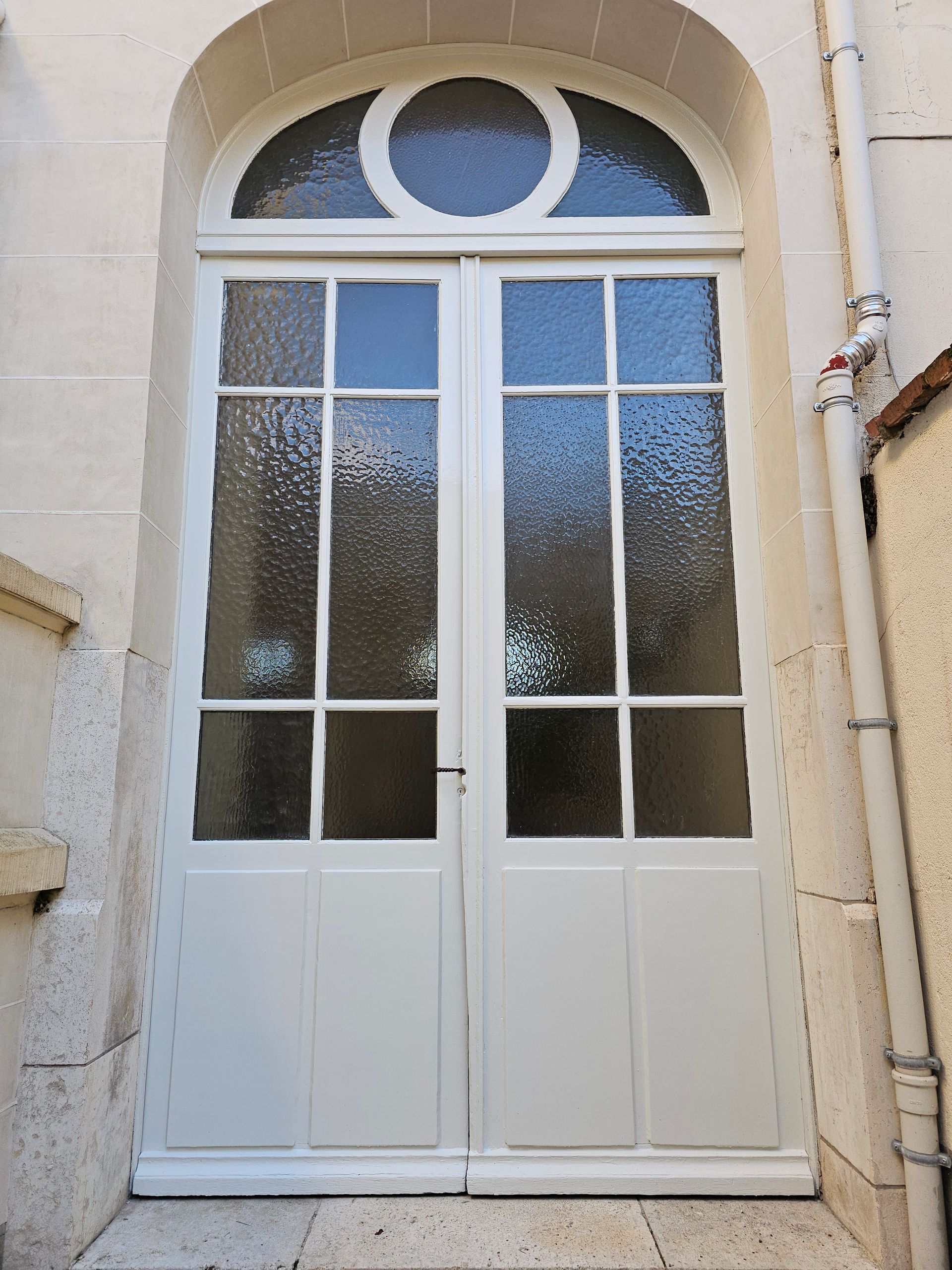Double porte cintrée blanche avec panneaux de verre et fenêtre circulaire au-dessus.