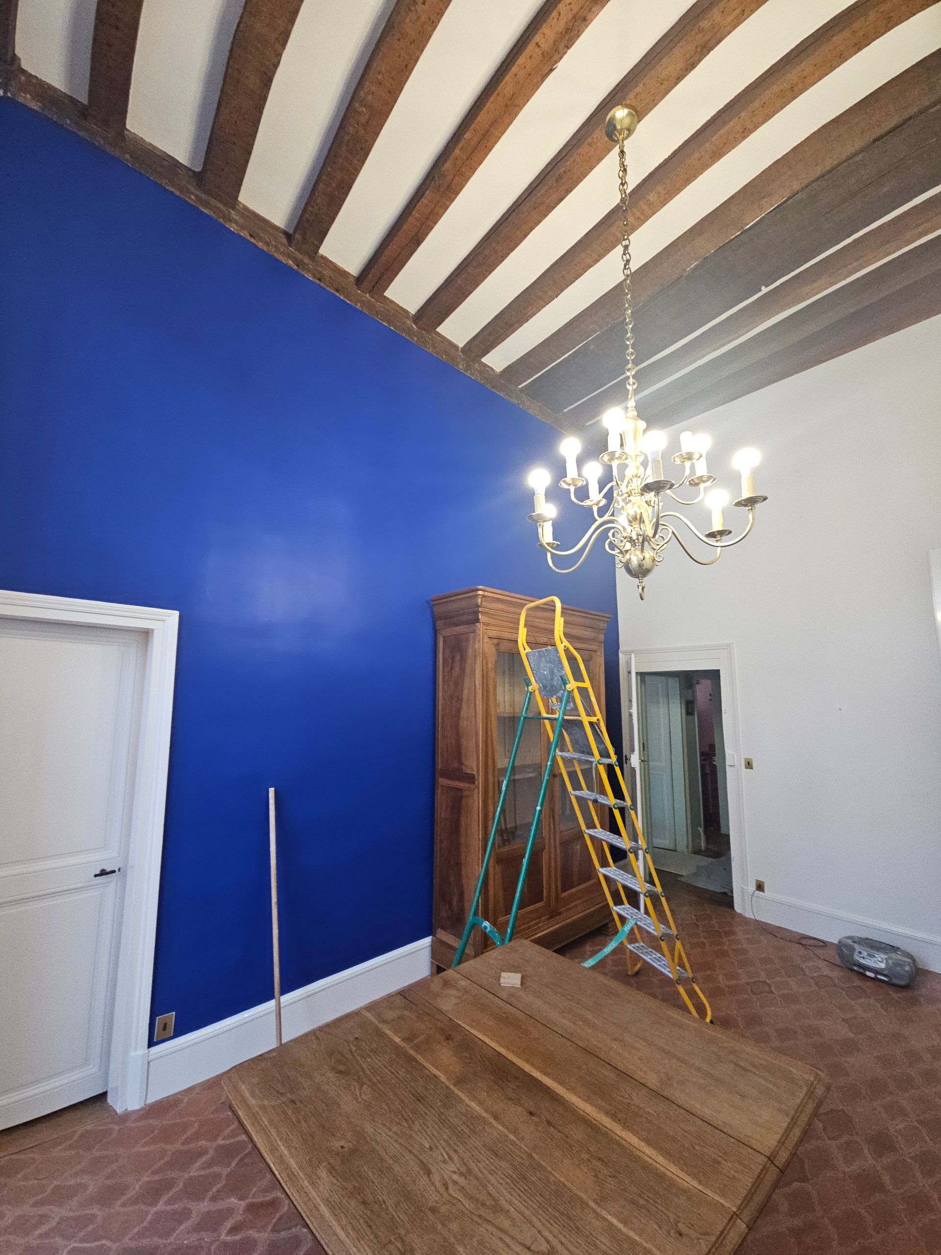 Pièce avec mur bleu, poutres en bois, lustre, armoire, échelle et table.