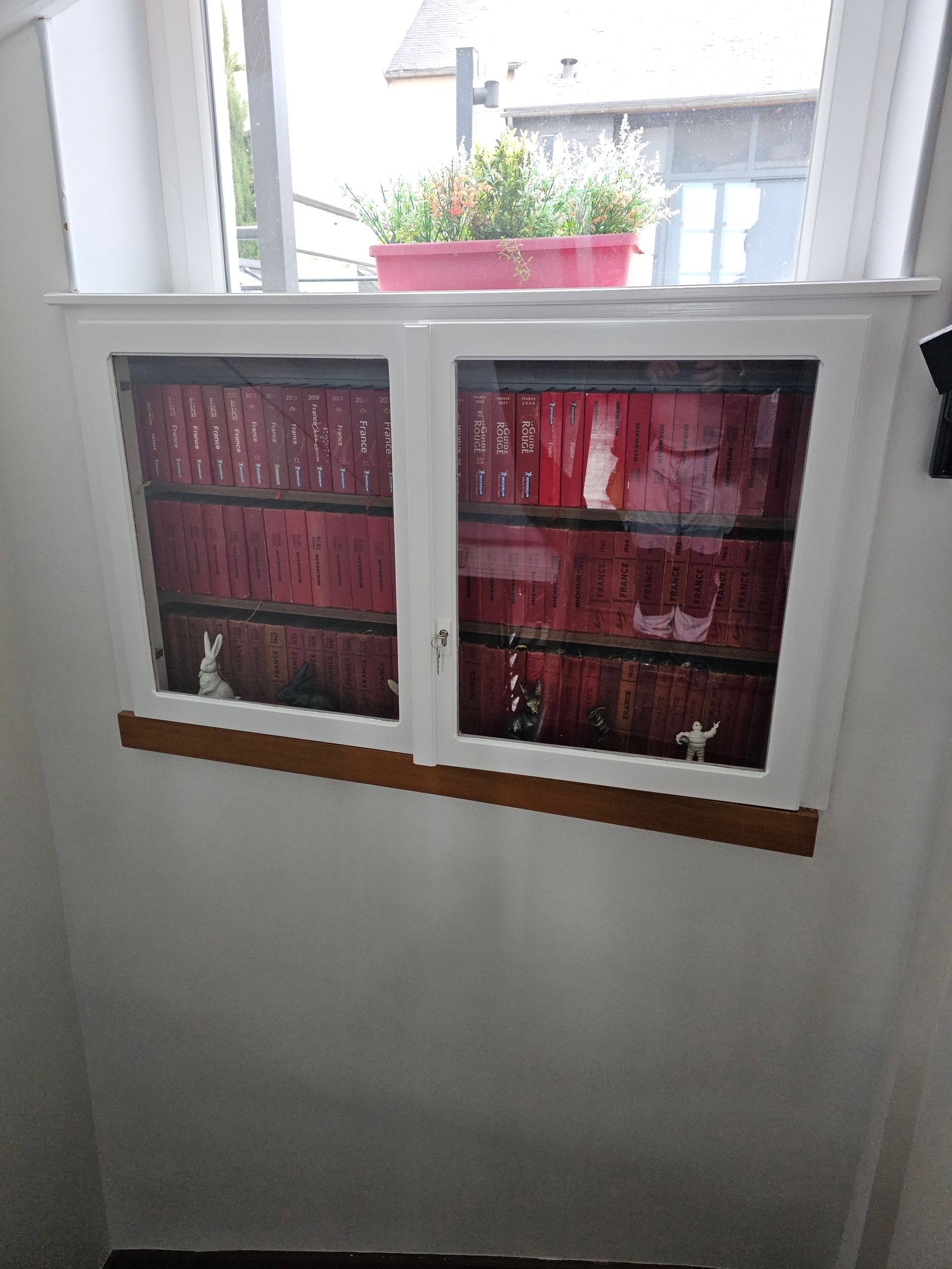 Fenêtre ornée d'une reproduction d'une étagère à livres rouges, encadrée de blanc, avec une petite jardinière posée dessus.