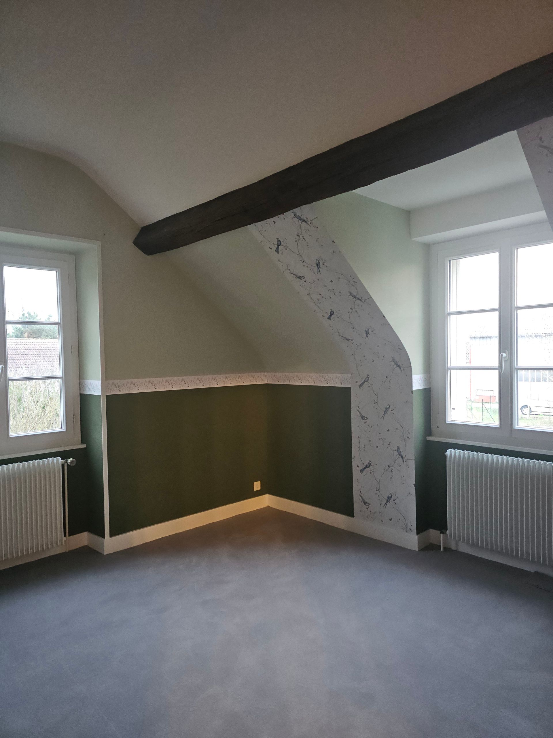 Pièce avec deux fenêtres, murs verts et blancs, poutre sombre et sol gris.