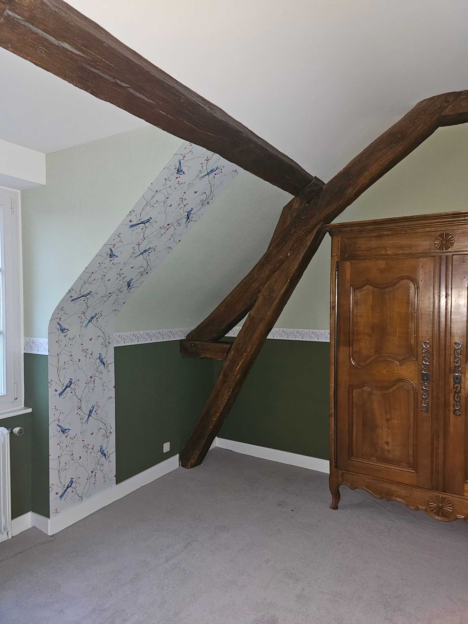 Chambre avec poutres apparentes, murs vert pâle, papier peint fleuri bleu et armoire en bois.