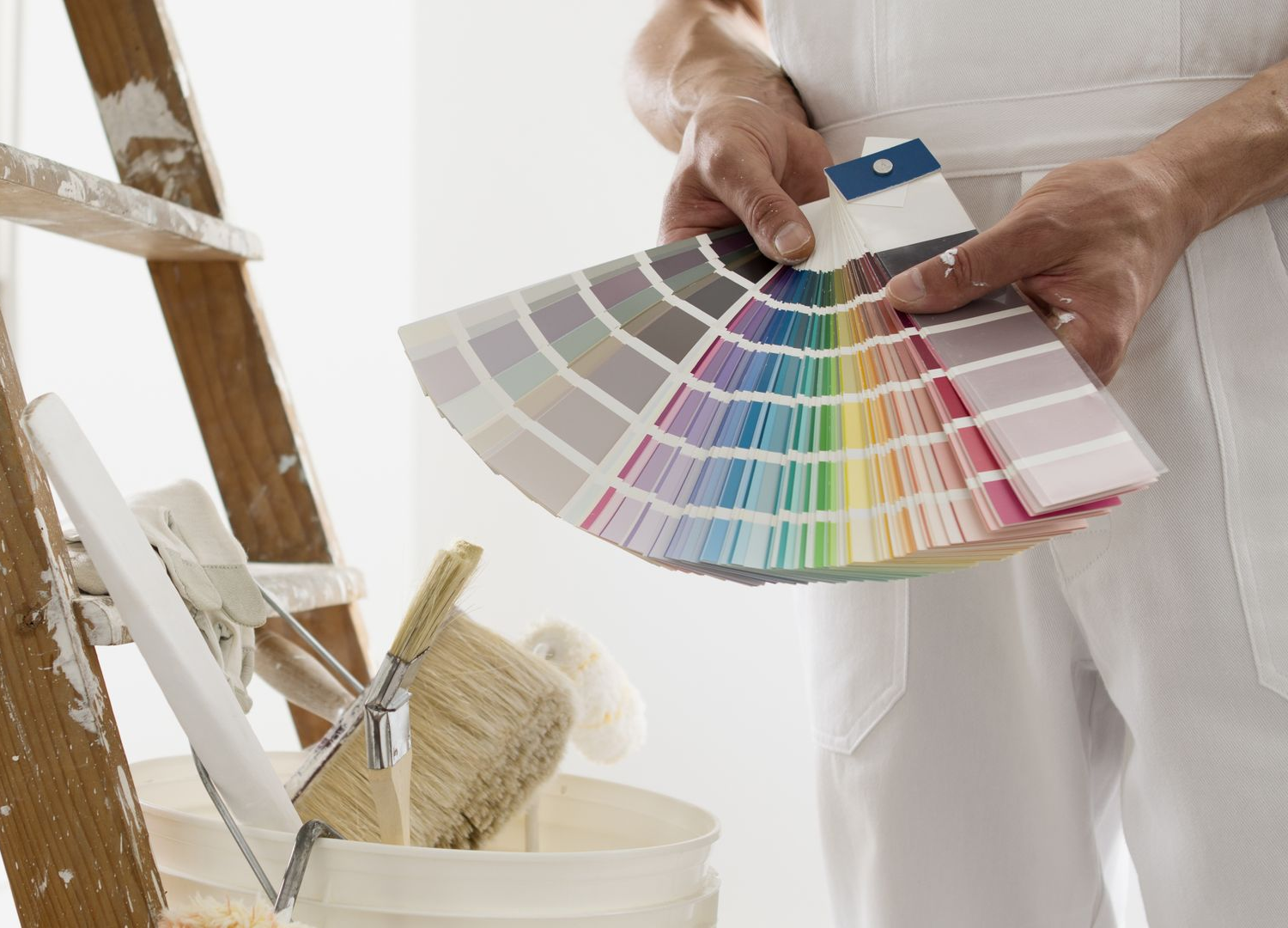 Une personne en salopette blanche tient un éventail d'échantillons de peinture près d'une échelle et d'un seau de peinture.