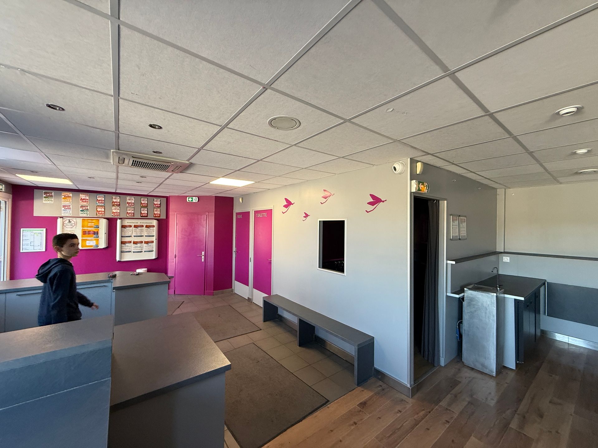 Une personne se tient debout dans un intérieur moderne avec des comptoirs gris, des portes roses et un banc.