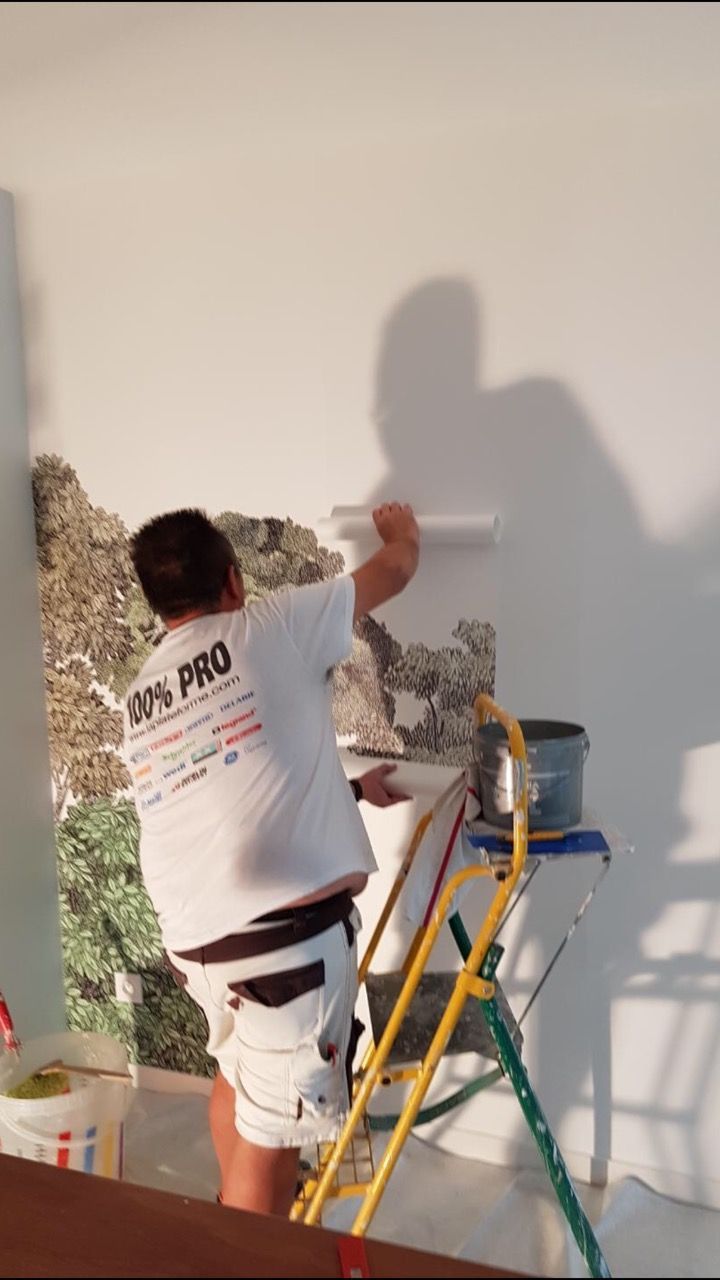 Une personne applique du papier peint au rouleau sur un mur, debout sur une échelle.