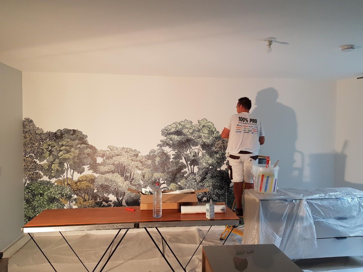Une personne peignant une fresque murale représentant une scène de forêt, debout sur une table dans une pièce.