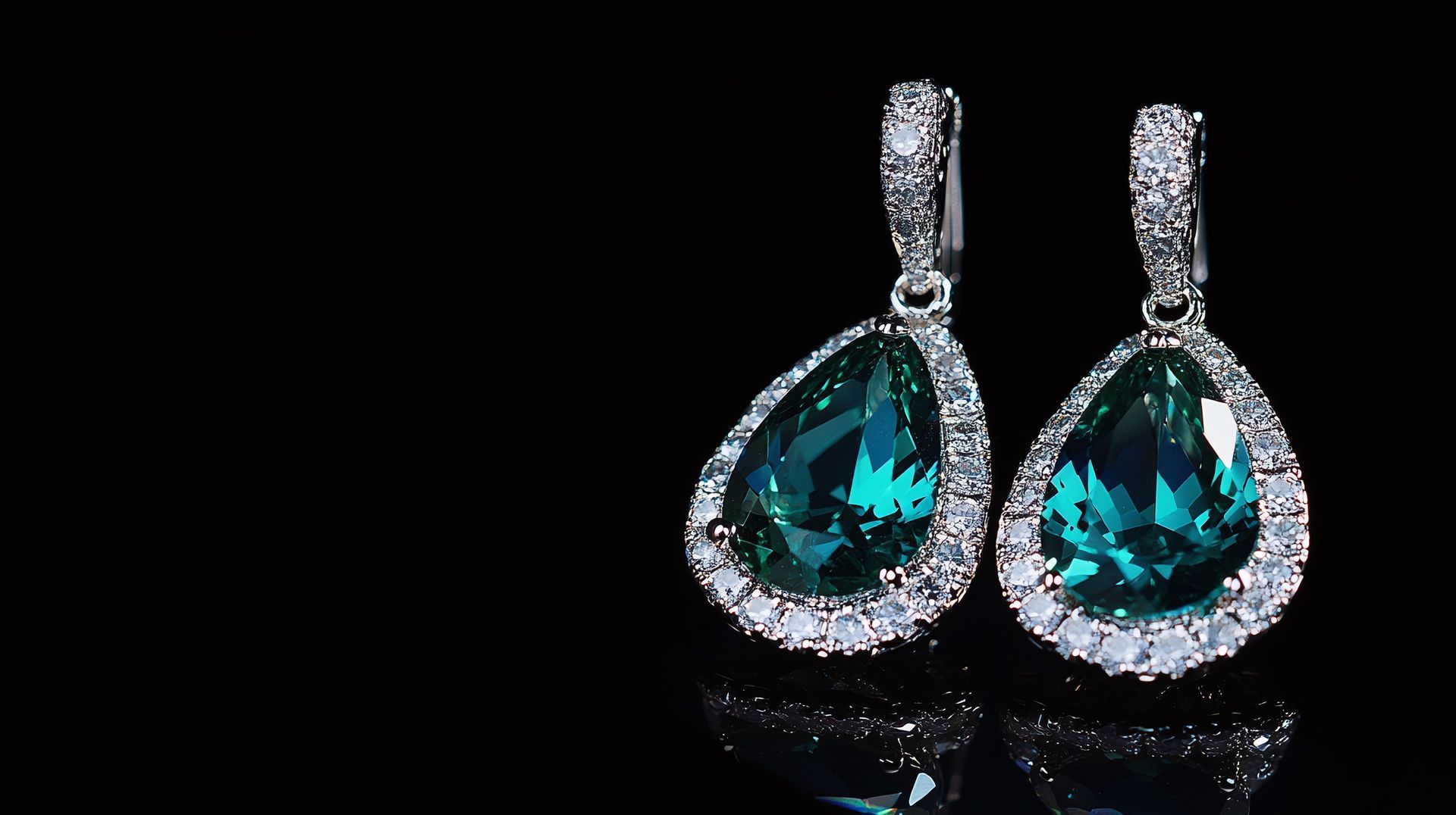 Ein Paar silberne Tropfenohrringe mit großen, tropfenförmigen, blaugrünen Steinen, umgeben von kleinen, klaren Edelsteinen auf einer schwarzen Oberfläche.