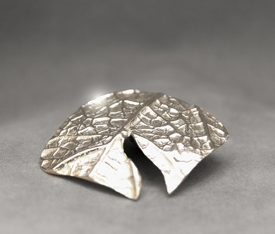 Silberne Brosche in Blattform mit strukturierter Oberfläche auf grauem Hintergrund.