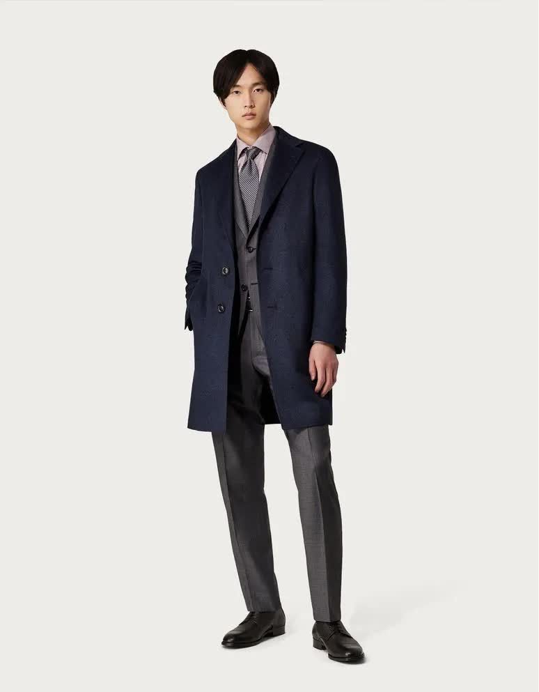 Homme en manteau bleu marine, costume et chemise à motifs, posant devant un fond blanc.