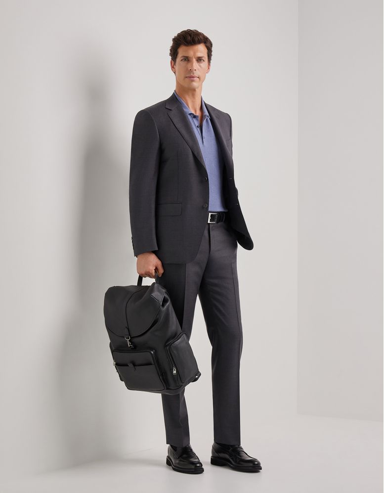 Un homme en costume sombre, debout contre un mur clair, tenant un sac à dos noir.