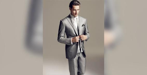 Un homme en costume gris, chemise blanche et cravate, avec une pochette, pose en studio.