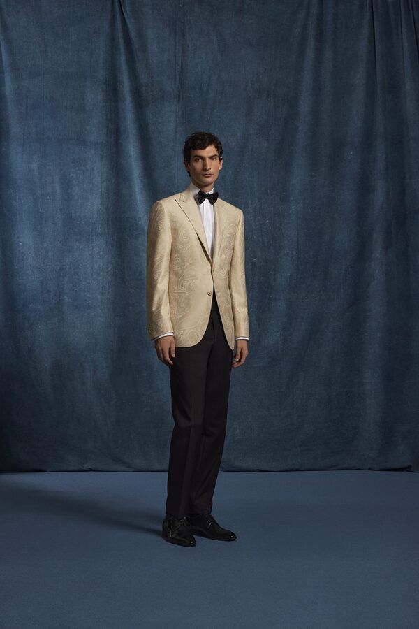 Homme en blazer crème, pantalon noir et nœud papillon, se détachant sur un fond bleu.