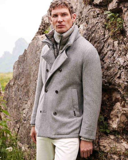 Un homme vêtu d'un manteau gris croisé et d'un pantalon blanc se tient devant une formation rocheuse.