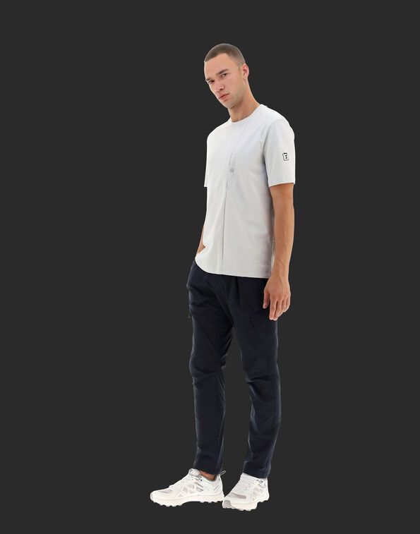 Homme en chemise gris clair et pantalon bleu marine, debout, regardant la caméra.