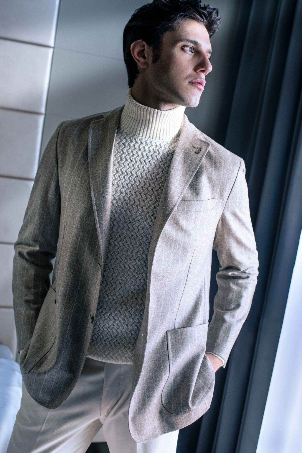 Un homme vêtu d'un blazer beige clair et d'un pull à col roulé, debout près d'une fenêtre, regarde au loin.