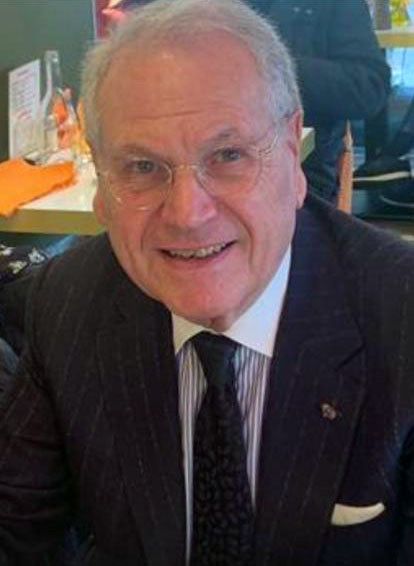 Un homme portant des lunettes, un costume et une cravate, sourit à la caméra.