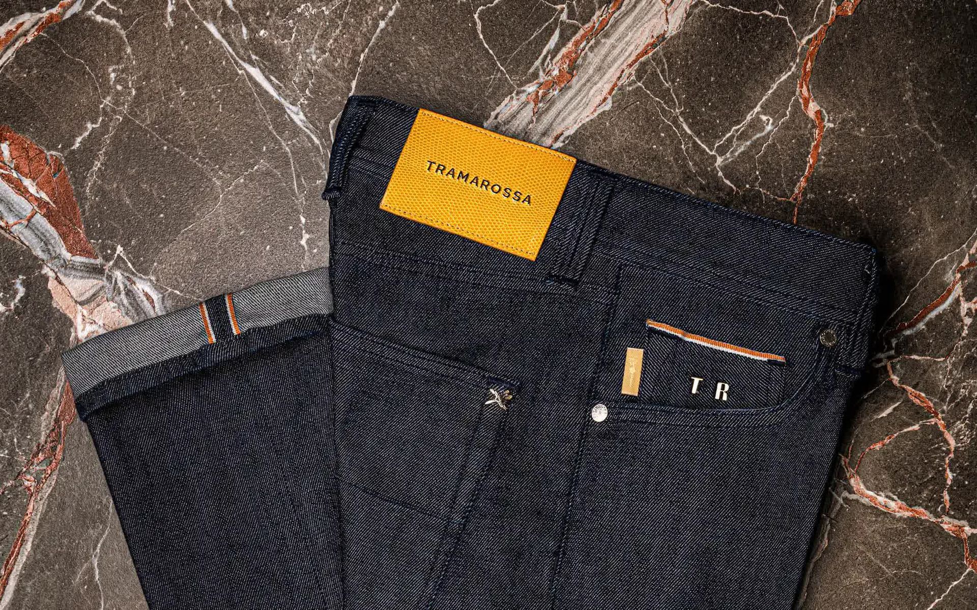 Jean en denim foncé avec surpiqûres orange et étiquette jaune sur une surface en marbre.