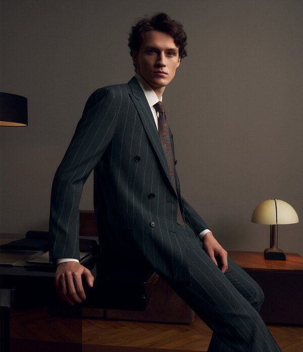 Un homme en costume vert à fines rayures, appuyé sur un bureau, regarde l'objectif. Ambiance sombre et mystérieuse.