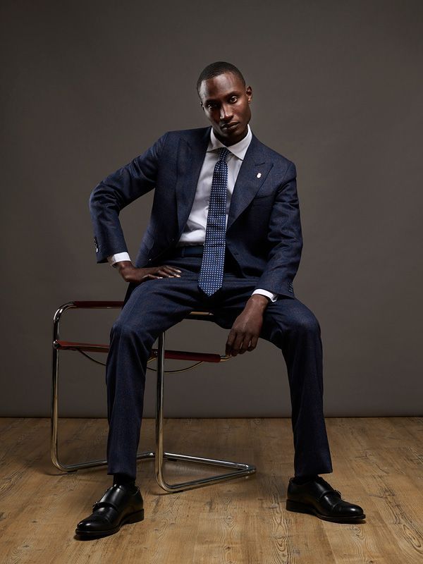 Un homme en costume bleu, assis sur une chaise en métal, regarde l'objectif. Il a une main sur la hanche.