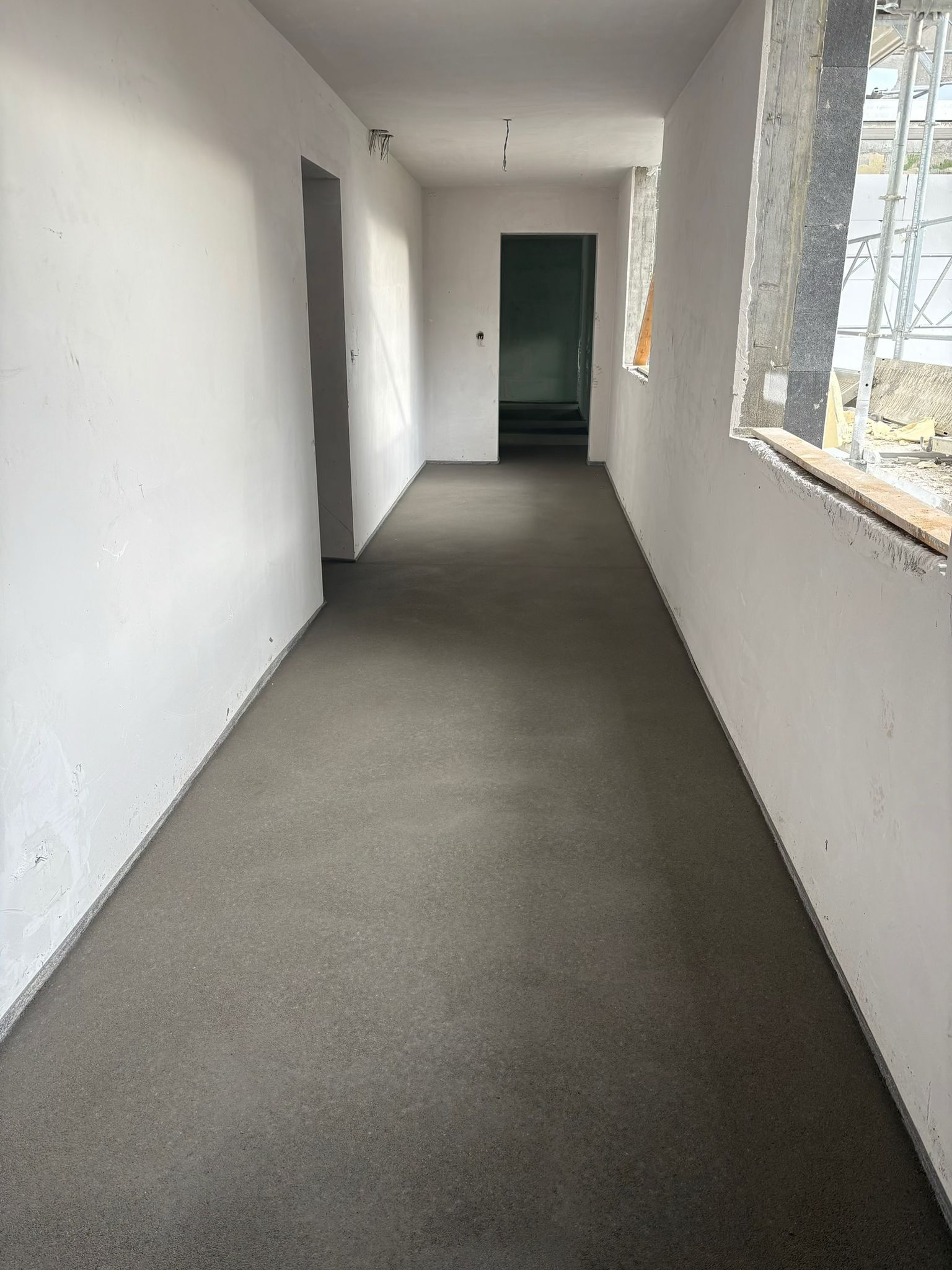 Un couloir en construction avec des murs en plâtre blanc, un sol en béton gris et deux portes donnant sur des pièces sombres.