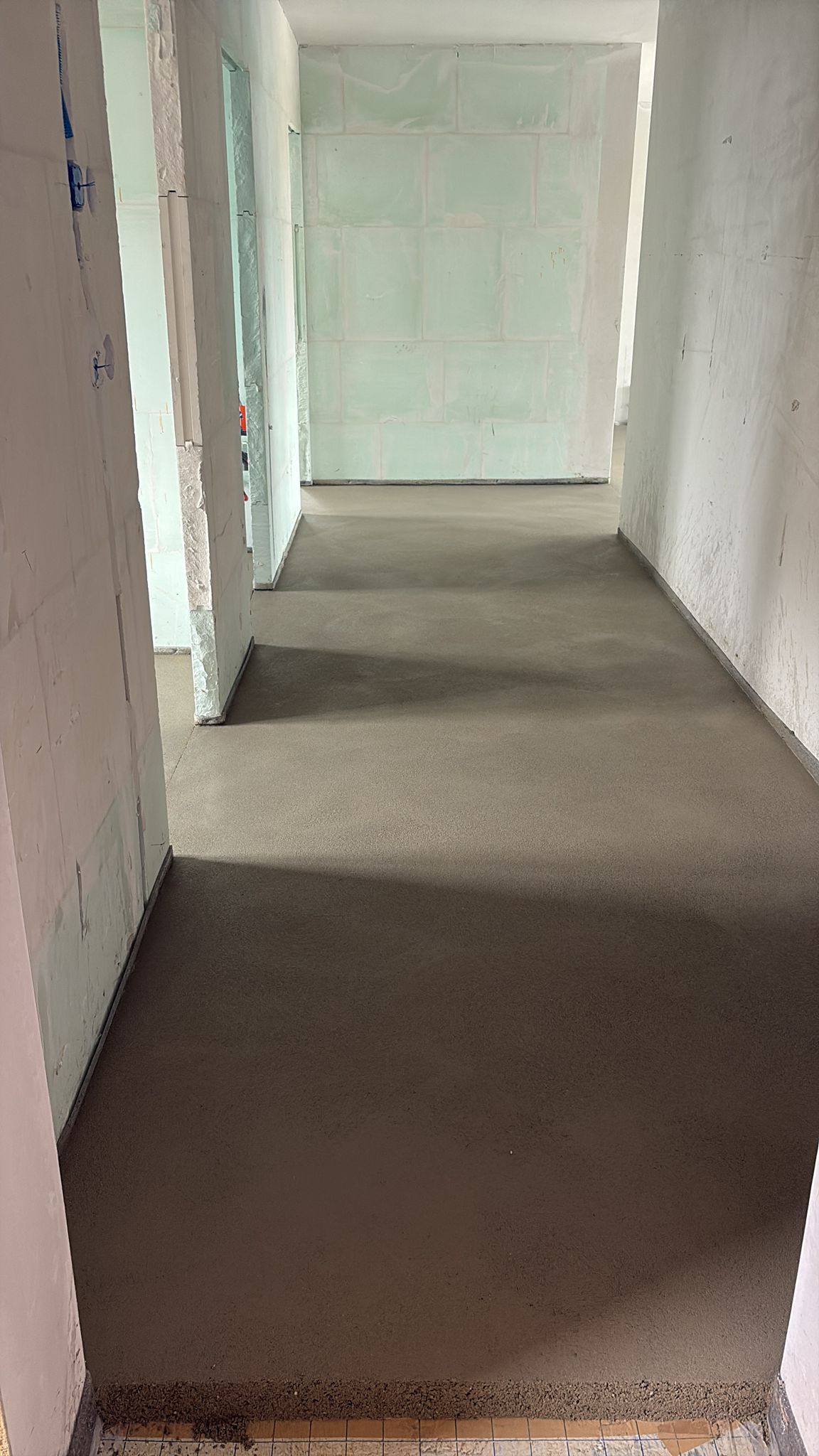 Un couloir en construction avec un sol en béton gris fraîchement coulé, des murs blancs non finis et des portes apparentes.