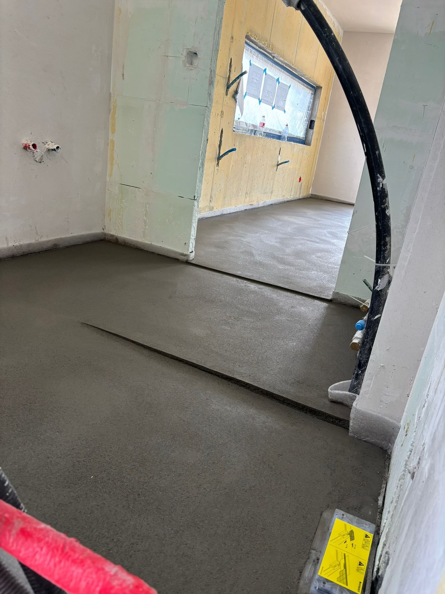 Une pièce en construction avec un sol en béton fraîchement coulé, de l'isolant apparent et un tuyau noir dressé à la verticale.