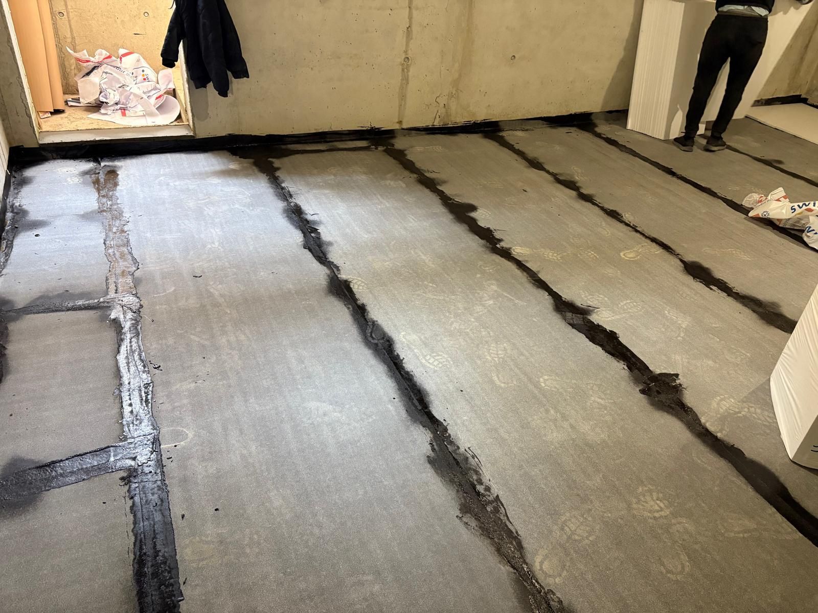 Dans une pièce en rénovation, le sol est recouvert de bandes de sous-couche grises, séparées par de larges joints adhésifs foncés.