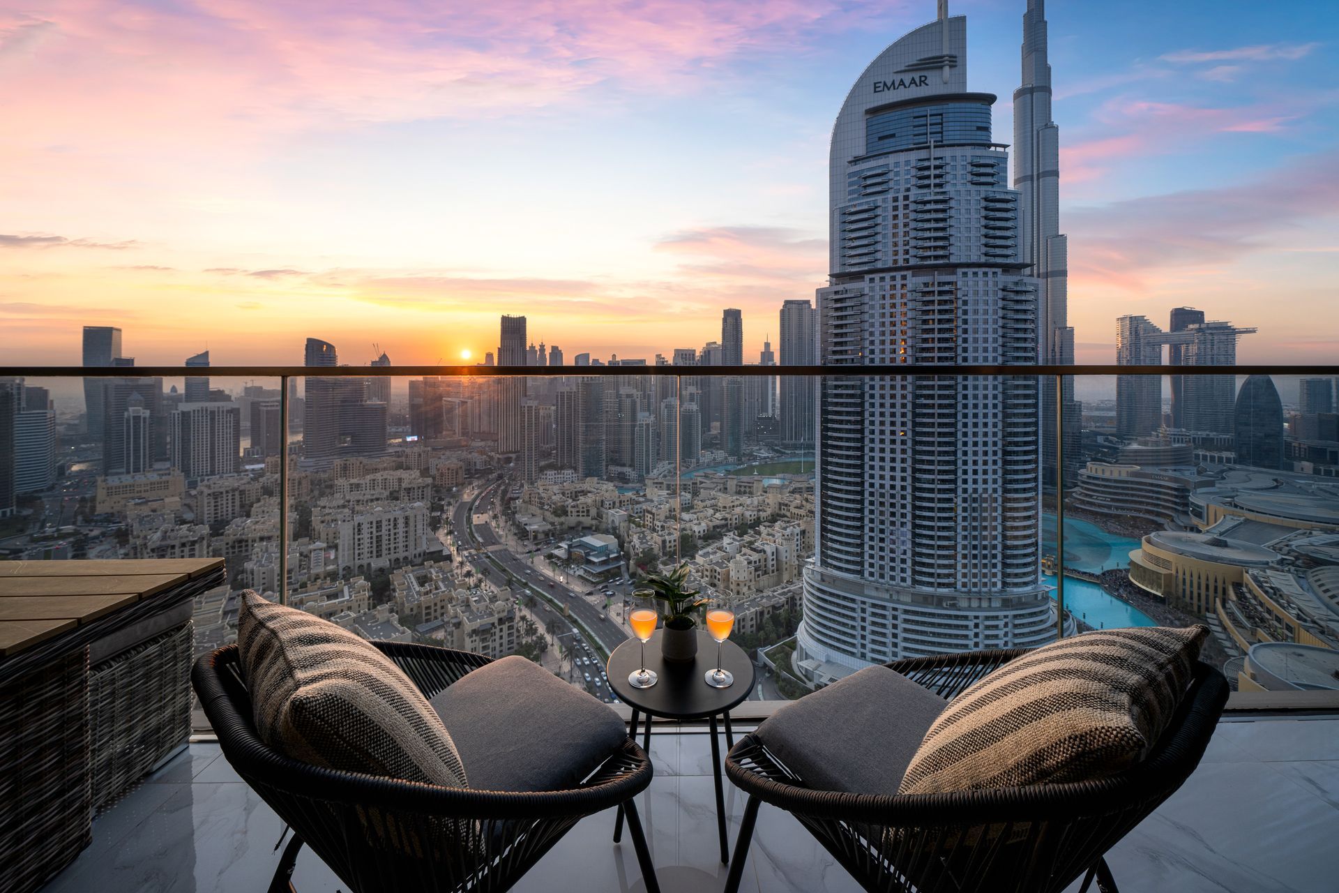 Balcony view of Dubai skyline at sunset, with chairs, small table, drinks, and the Burj Khalifa.