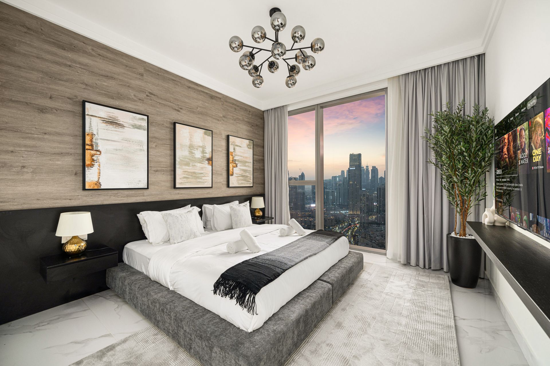 Modern bedroom with city view, gray tones, artwork, large bed, and chandelier.