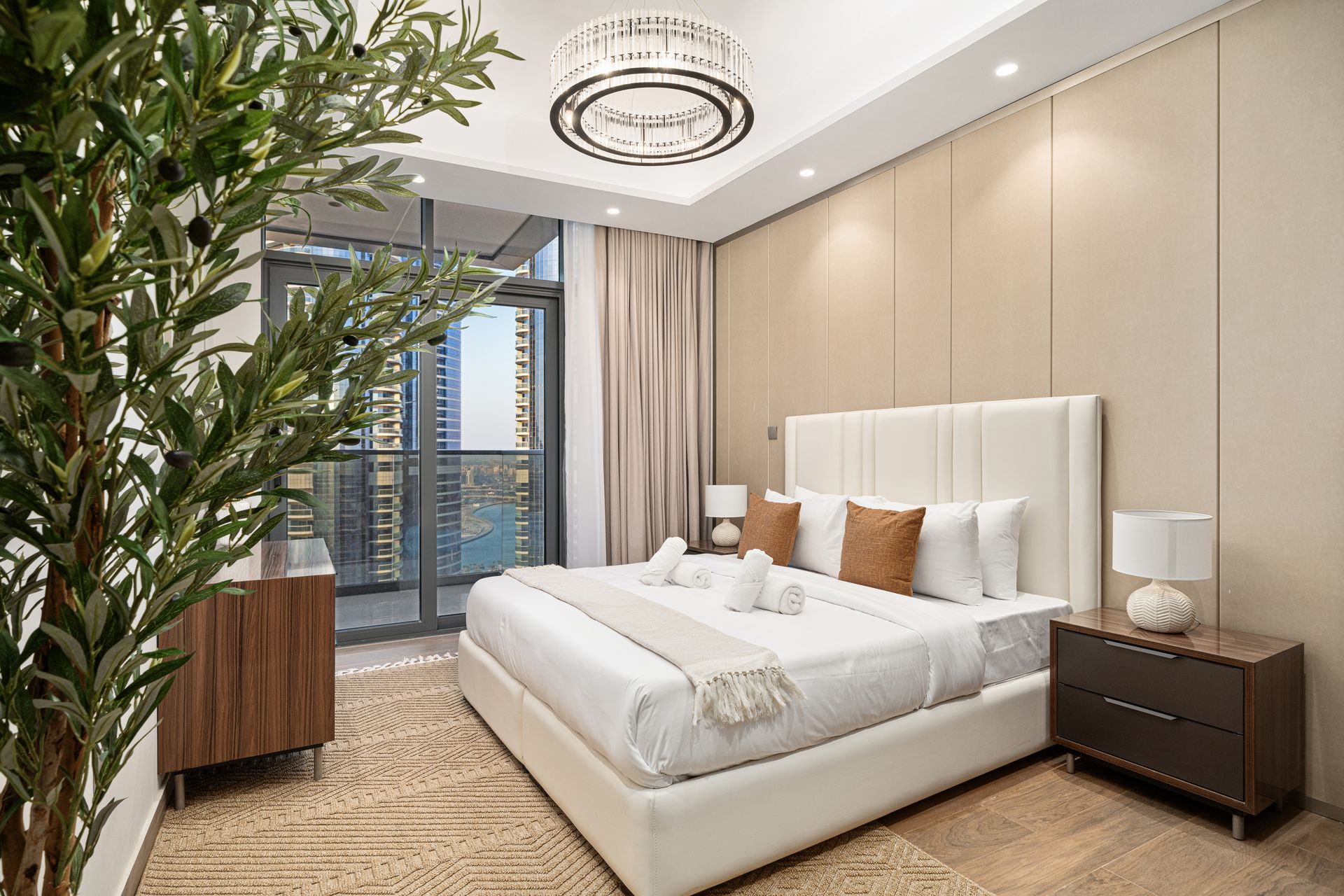 A modern bedroom with a large bed, tall window, and city view, neutral tones.