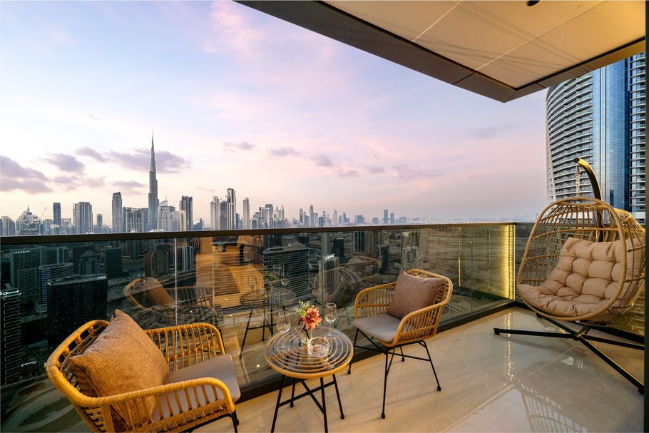 Balcony with city view: wicker chairs, hanging chair, glass table, cityscape at sunset.