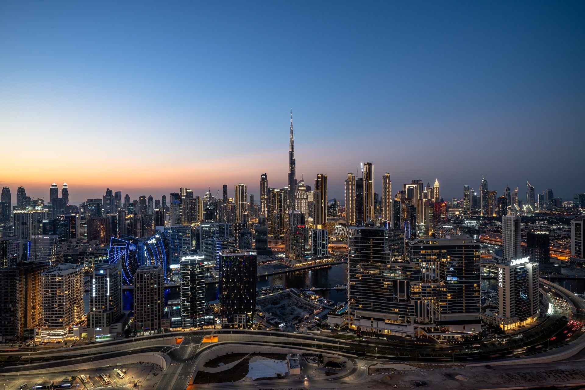 Dubai skyline at dusk with the Burj Khalifa illuminated, buildings lit up, and a calm blue sky.