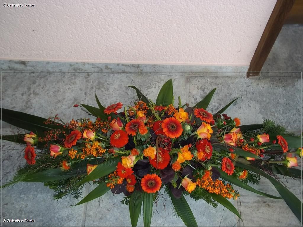 Langes Trauergesteck mit orangefarbenen Blüten, grünen Blättern und strukturierter Form.