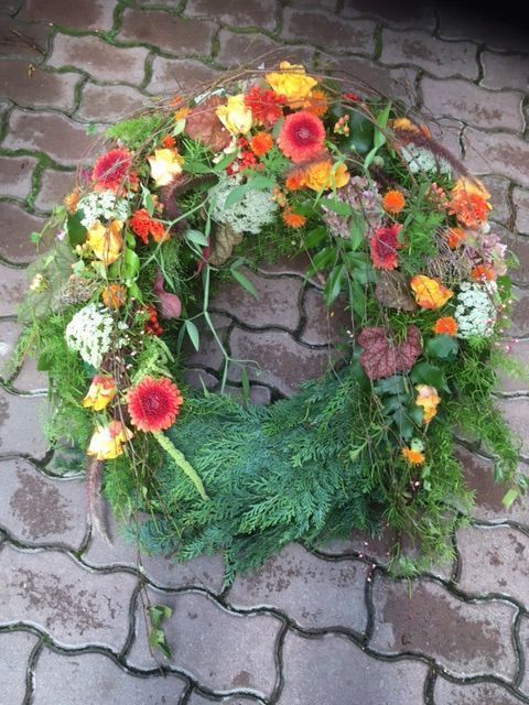 Farbenfroher Trauerkranz mit orangefarbenen Gerbera, gelben Blüten und saisonalem Grün.