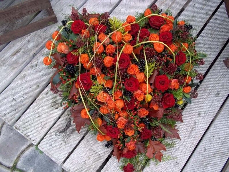 Herzförmiges Trauergesteck mit orangefarbenen Rosen und grünem Blattwerk.