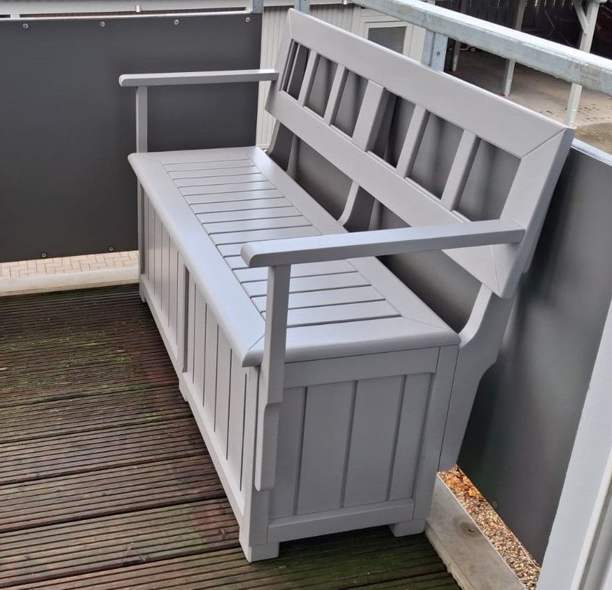 Graue Holzbank mit Stauraum auf einem Deck, mit Armlehnen und Rückenlehne.