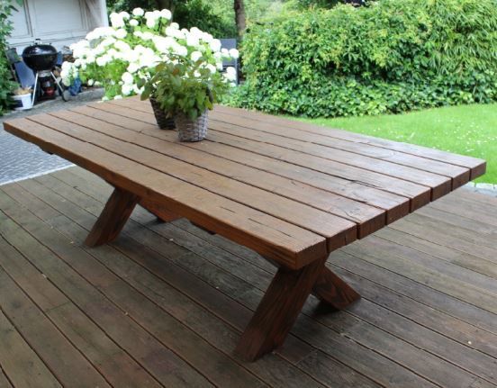 Hölzerner Gartentisch mit X-förmigen Beinen, darauf zwei Topfpflanzen. Im Hintergrund ist eine Terrasse zu sehen.