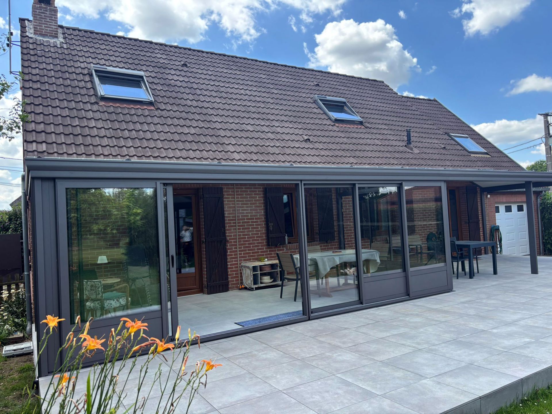 Ajout d'une véranda à une maison en briques avec de grandes portes vitrées, un cadre gris et un patio carrelé.