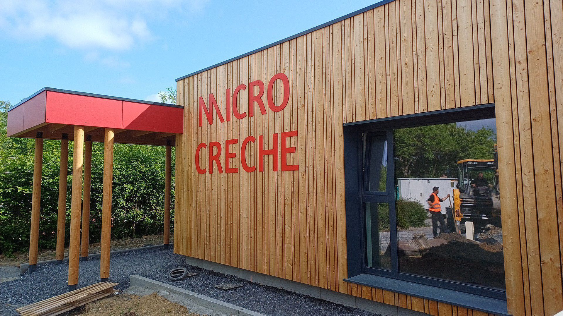 Bâtiment en bois avec l'inscription « MICRO CRÈCHE » en lettres rouges. Auvent rouge. Homme en gilet orange visible par la fenêtre.