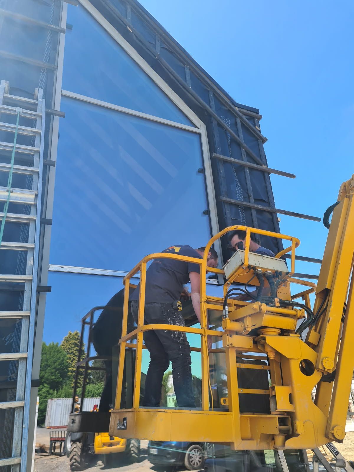 Des ouvriers du bâtiment installent une grande fenêtre sur un bâtiment à l'aide d'un ascenseur jaune sous un ciel bleu.