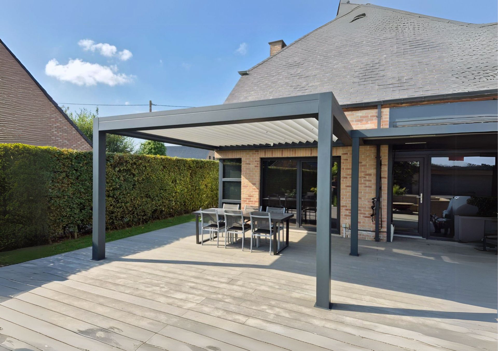 Pergola au dessus d'un patio avec table et chaises, à côté d'une maison en briques avec une haie verte.
