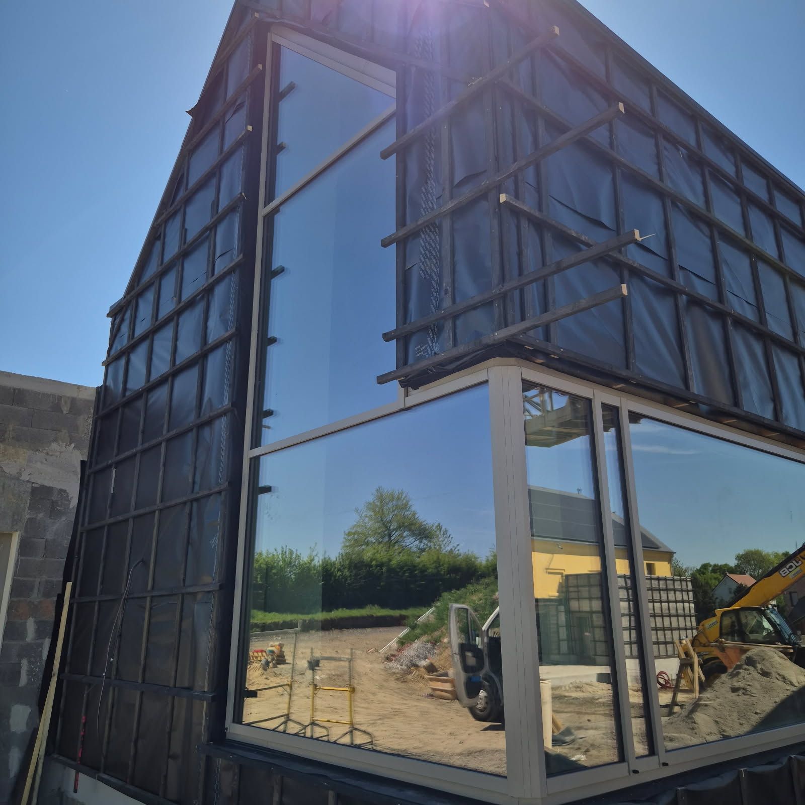 Maison à ossature en A en construction avec de grandes fenêtres reflétant l'environnement, y compris une excavatrice jaune.
