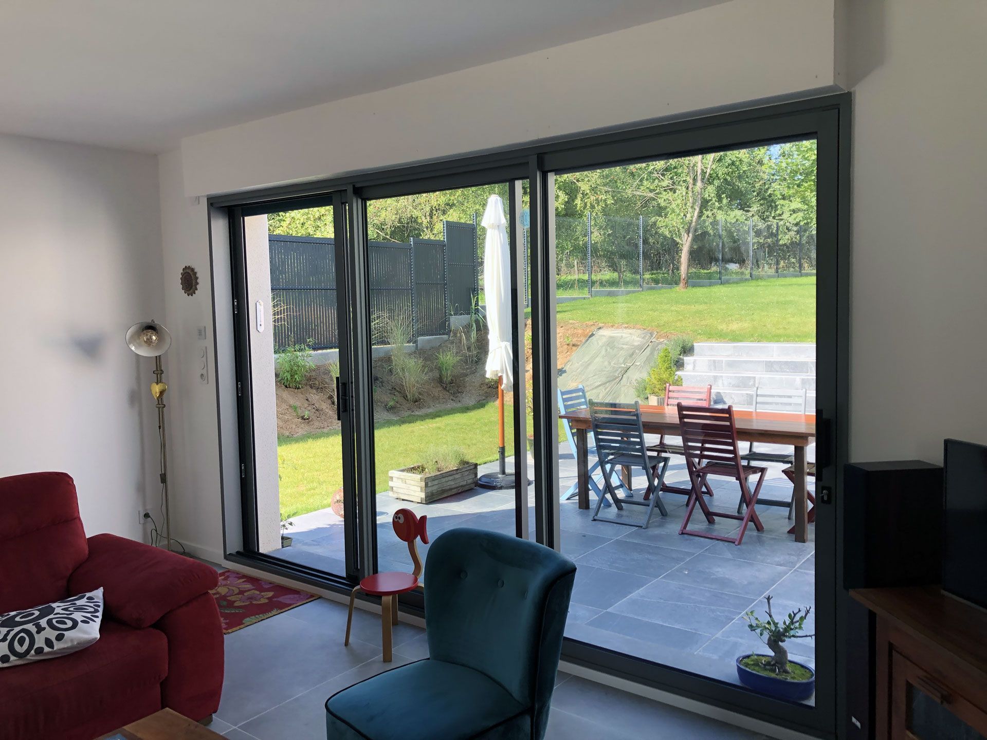 Salon avec de grandes portes coulissantes ouvertes sur un patio arrière, canapé rouge et chaise bleu sarcelle au premier plan.
