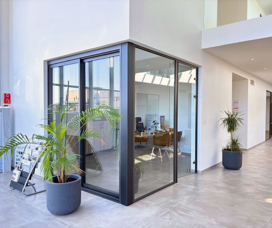 Espace de bureau vitré à l'intérieur d'un bâtiment moderne, plantes en pot devant, vue sur le mobilier à l'intérieur, lumière vive.